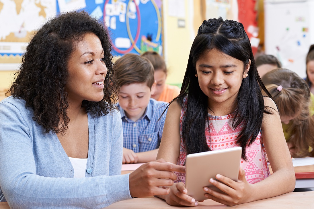 Teacher Helping Group Of Elementary School Children In Computer